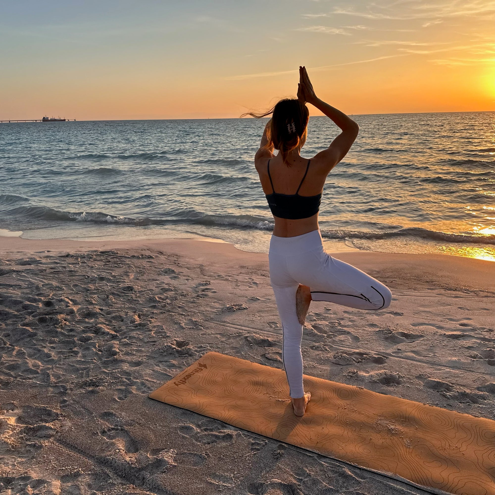 Fitnexfly donna in equilibrio su una gamba con l'altra a 90 gradi vista di schiena che saluta il sole al tramonto
